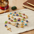 A rosary necklace with colorful Turkish evil eye beads and a gold cross rests on beige cloth beside a notebook, pen, incense, and green stone.